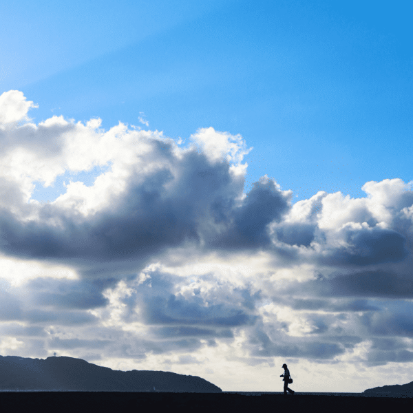 A person walks under a sky of fluffy clouds, embodying the art print Sky, with hills silhouetted on the horizon and sunlight streaming through.