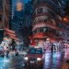 A red taxi glides through a rain-soaked street in a bustling city at night, resembling an Amber Rain art print. Neon lights illuminate the wet pavement as tall buildings rise in the background, while pedestrians with umbrellas stroll along the sidewalks.