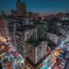 The Night Fall features a vibrant night market scene like an art print, with colorful street stalls lit brightly amidst tall residential buildings under a dark sky, set against a city skyline at dusk.