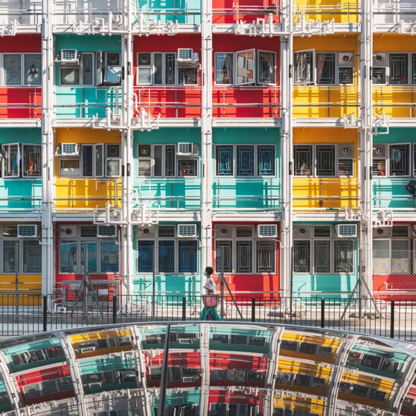 The Rainbow Apartment displays rows of windows in red, teal, and yellow. Below, a woman walks by a curved mirrored surface reflecting the structure. The vibrant scene resembles an art print, beautifully capturing dynamic urban life and architecture.