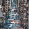 A busy urban street scene from Rooftop in Mong Kok captures Hong Kong with vehicles like buses and vans amid towering buildings. Vibrant Chinese signs add color as throngs of people cross, showcasing the citys bustling energy reminiscent of an art print.