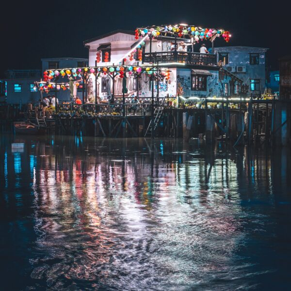 The Tai O Stilt House art print captures a waterfront house illuminated by colorful lanterns reflecting in the water at night, creating a festive atmosphere against the dark sky with gentle ripples distorting its mirrored image.