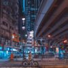 A cyclist stands with a bike at a city intersection at night, resembling an art print. The scene features a large overpass and tall buildings with bright neon signs. The cityscape glows in blue and orange tones, creating a vibrant urban atmosphere. Product Name: Cyclist.