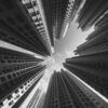 The Urban Jungle black and white art print showcases towering skyscrapers from a ground view, forming a circular pattern against a cloudy sky, offering a dramatic perspective with converging geometric lines.