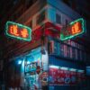 A night scene of a street corner showcases a building adorned with neon Chinese characters, evoking an art print. The green and red signs cast vivid hues. Below, a brightly lit storefront presents tempting food images. The classic urban look embodies the charm of Elderly.