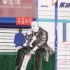 A bald man in a coat sits on a bench holding an object, flashing a peace sign. Behind him, someone in a blue jacket stands among Chinese and English signs, showcasing Telecom and Digital on the Simple Taste 11 art print.
