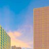 Sunset City captures a vibrant cityscape with sleek, grid-patterned buildings set against a blue sky filled with soft clouds and a pink-orange horizon, reminiscent of modern art.