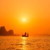 The Dragon Boat art print showcases a silhouette of a traditional sailing ship on an orange sea at sunset, with the sun setting behind city skyscrapers on the left, casting vibrant orange reflections against the sky and water.