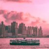 Under a vibrant pink and orange sky, the Ferry in Motion glides across the water with a city skyline and tall buildings in the background, all enhanced by fluffy clouds decorating the colorful sunset.