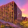 In Unclear Vision, a digitally enhanced urban scene emerges with tall, reflective buildings at sunset casting warm pink, orange, and purple hues. Lush greenery beautifully contrasts the modern architecture in this captivating art print.