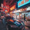 A vibrant city street at night, as depicted in Portland Street, showcases bright neon signs in various languages. A black car is parked on the roadside while a person walks past a row of illuminated shops with colorful signs and advertisements.