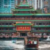Jumbo Kingdom, a large, ornate floating restaurant with traditional Asian architecture in red and green hues, graces the water like an art print. Two boats navigate the waterway in front of the urban skyline.