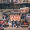 A small, cluttered shop named Master Wong offers a variety of goods and colorful textiles. Two people stand among vibrant art prints. Chinese signage above the shop shows phone numbers, and a balcony with railing is visible above.