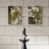 A textured tiled wall features two abstract Sunset Shadow art prints with swirling patterns, while below, a small marble pedestal with a smooth dark finish holds a metal knights helmet.
