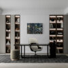 A modern office features a sleek black desk, a beige chair on a patterned rug, and two illuminated bookshelves displaying books, vases, and sculptures. An art print titled Chromatic Convergence is split and hangs on the wall between the shelves against a neutral backdrop.