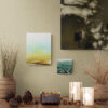 A warm and tranquil setting features the iooooi tabletop scene with two metallic candle holders, pinecones, potted grass, and three earthy-toned abstract art prints on the wall.